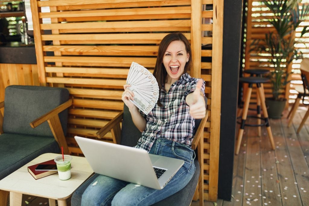 Woman in outdoors street coffee shop cafe sitting with modern laptop pc computer hold in hand bunch of dollars banknotes cash money Mobile Office restaurant in free time Freelance business concept